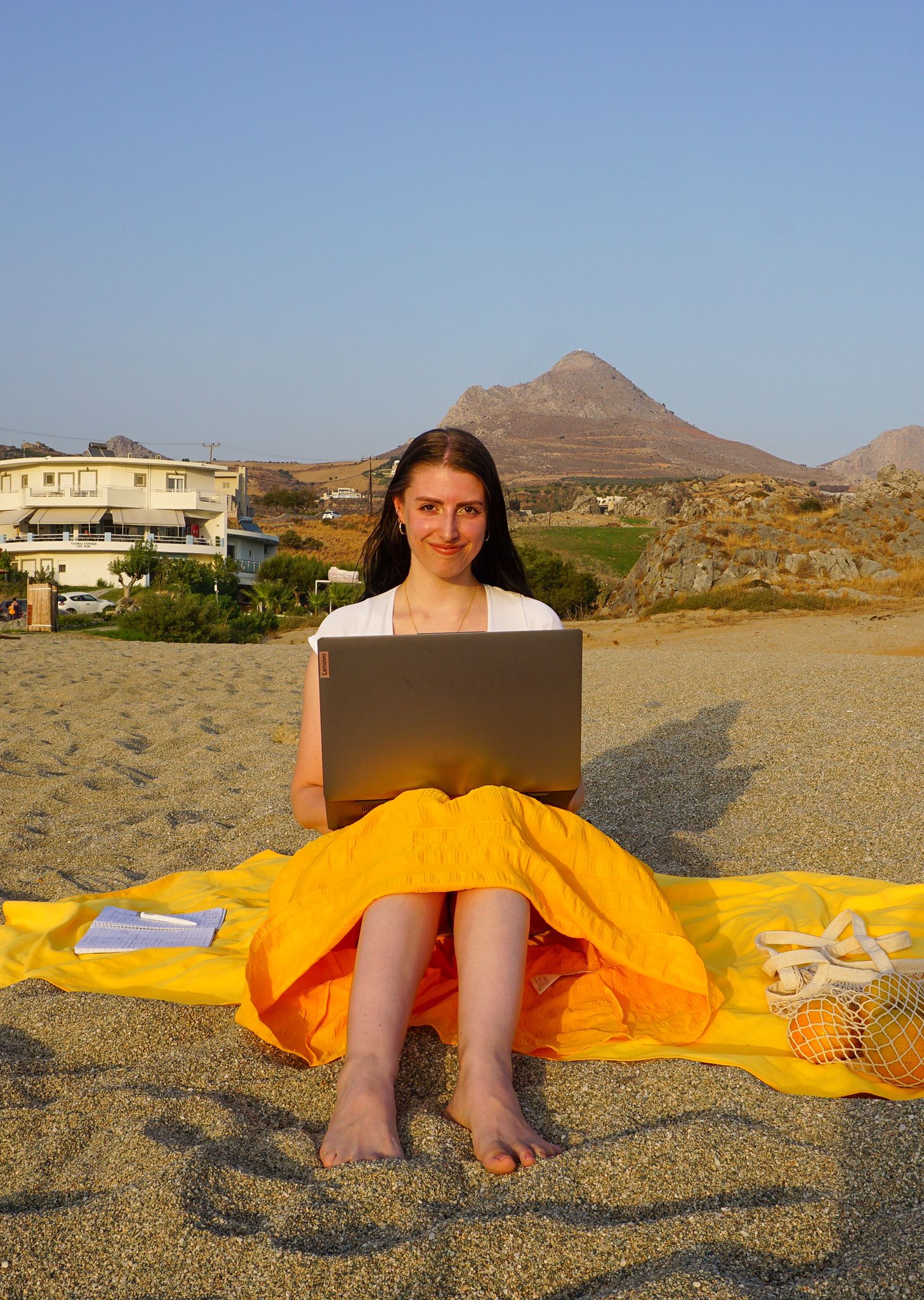 Anki sitzt mit Laptop am Strand, im Hintergrund sind ein Haus und Berge